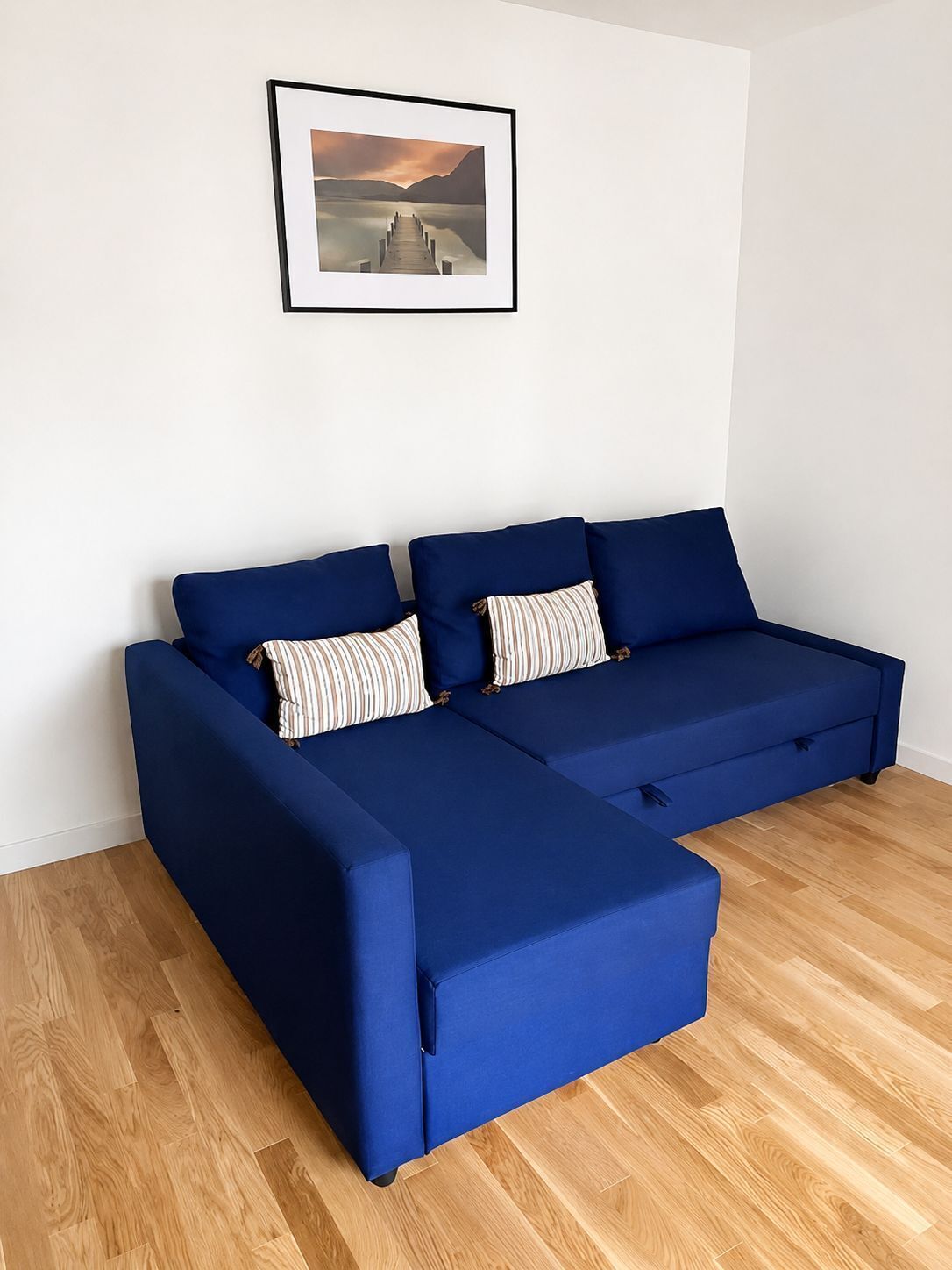 Ein blaues Ecksofa mit gestreiften Kissen, ein gerahmtes Bild darüber und Holzboden.