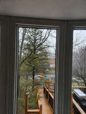 Näkymä ikkunasta, josta avautuu puinen terassi, joka johtaa leikkikentälle.
