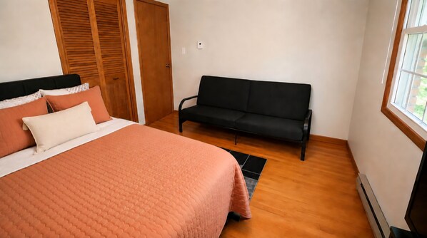 A bedroom with a bed, a sofa, wooden flooring, a window, and a wardrobe.