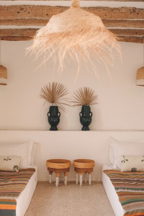 Une pièce comprenant un lit, deux petits tabourets en bois, une étagère fixée au mur avec deux vases décoratifs et un plafond recouvert de chaume.