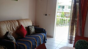 Sala de estar com sofá, mesa de jantar e planta em vaso, e vista para uma varanda através de uma porta de vidro.