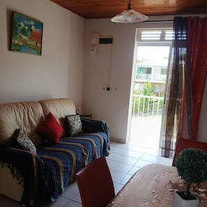 A living room with a sofa, a dining table with a potted plant, and a view of a balcony through a glass door. - L'ARUM 97280 (Le Vauclin)