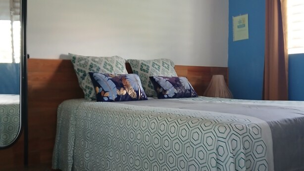Quarto com cama, teto de madeira, parede com detalhes em azul, janela com cortinas e espelho.