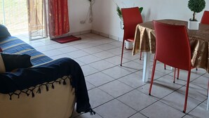 Quarto com cama, mesa de jantar com cadeiras, TV fixada na parede e um pequeno vaso com planta.