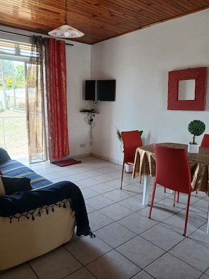 A room with a bed, a dining table with chairs, a TV mounted on the wall, and a small potted plant. - L'ARUM 97280 (Le Vauclin)
