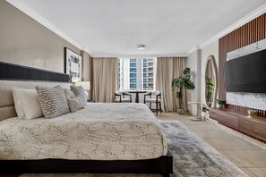 A modern hotel room with a large bed, a TV, a dining table, and a view of buildings. - Stylish Ocean view/front Studio + Pool (Sunny Isles Beach)