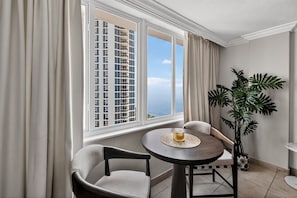 A room with a view of a tall building, a round table with a glass, two chairs, a potted plant, and a window with curtains. - Stylish Ocean view/front Studio + Pool (Sunny Isles Beach)