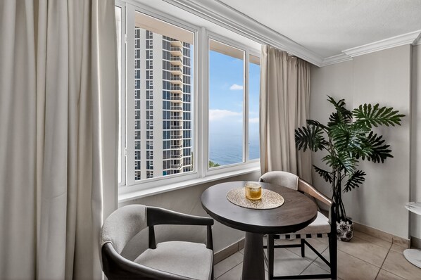 A room with a view of a tall building, a round table with a glass, two chairs, a potted plant, and a window with curtains. - Stylish Ocean view/front Studio + Pool (Sunny Isles Beach)