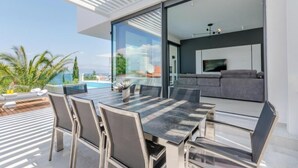 A modern outdoor dining area with a glass table and chairs, a pool, and a view of the ocean. - Villa Pax. (Sutivan)