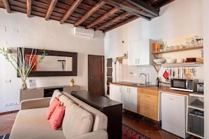 A modern kitchen with white cabinets, a microwave, and a sink. There is a wooden ceiling and a sofa with cushions in the living area. - BQA San Crisogono Apartments Rome-Trastevere (Roma)