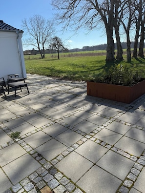 Un patio empedrado con zona de estar, una jardinera y vista a un campo cubierto de césped y árboles.