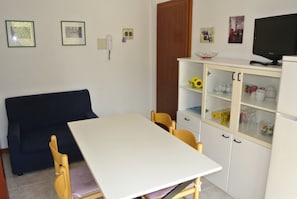 A room with a white table, two wooden chairs, a dark sofa, a white cabinet with glass doors, a television, and framed pictures on the wall. - Functional Flat Close to the Beach and the Center (Lignano Sabbiadoro)