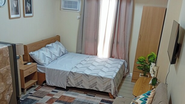 A bedroom with a bed, bedside table, lamp, air conditioning unit, curtains, a sofa, a small table, and a TV. - Cozy & Convenient | Cebu IT Park Stay (Cebu City)