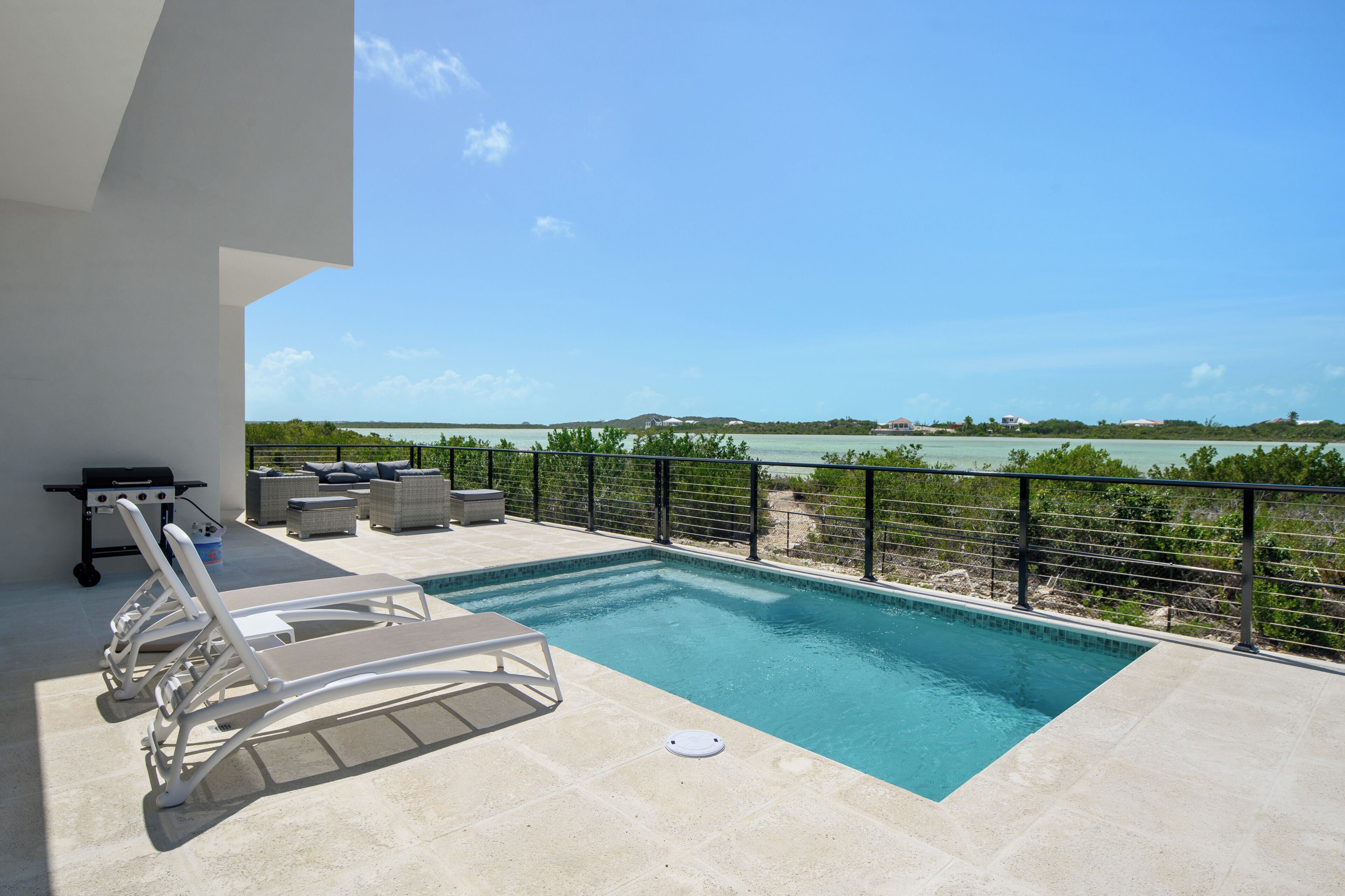 A pool with lounge chairs, a grill, and a view of the sea.