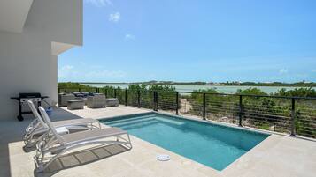 A pool with lounge chairs, a grill, and a view of the sea.