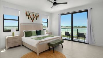 A bedroom with a bed, bedside tables, a ceiling fan, a view of the sea, and a balcony with chairs.