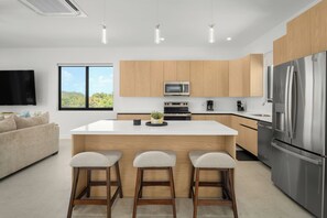 A modern kitchen with wooden cabinets, a central island with stools, a stainless steel refrigerator, and a flat-screen TV mounted on the wall. - Spacious 4-bedroom house with Gorgeous Views from Every Room (Venetian Road)