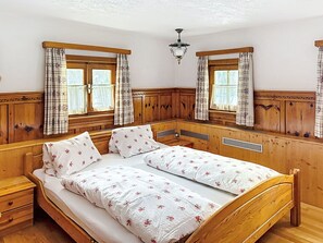 Una habitación de madera con una cama, dos ventanas con cortinas, una lámpara en el techo y un banco de madera.