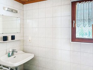 Un baño con azulejos blancos, un lavamanos, un espejo y una ventana con cortina estampada.