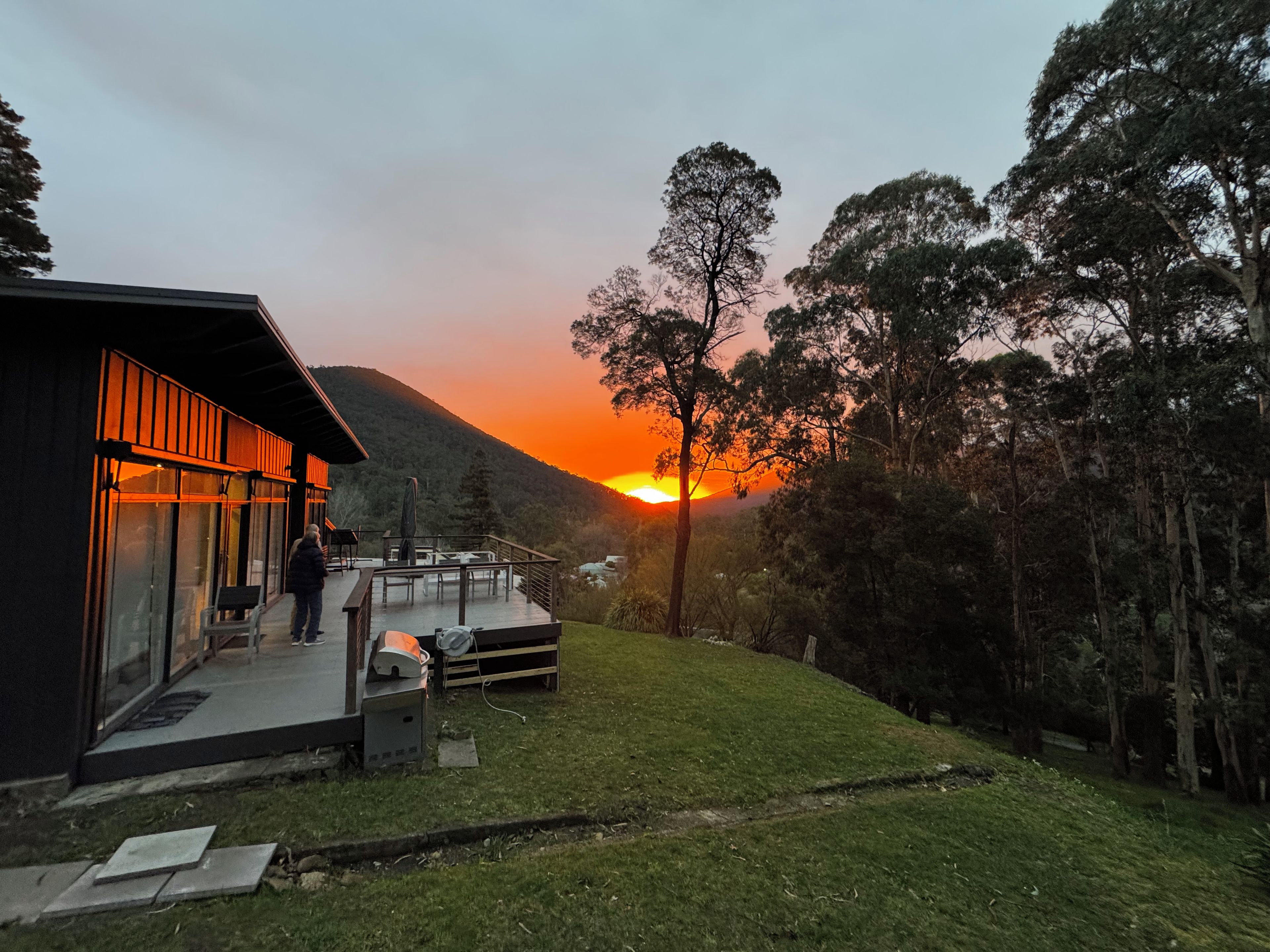 A modern building with a glass facade, a deck, and a view of the sunset over a hilly landscape.