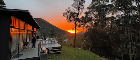 A modern building with a glass facade, a deck, and a view of the sunset over a hilly landscape.