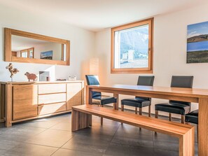 Ein modernes Zimmer mit einem Holztisch, Stühlen, einer Bank, einem großen Fenster mit Blick auf die Berge, einem Spiegel und einem Schrank.