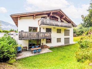 A two-story chalet with a wooden balcony, a patio with outdoor seating, and a mountain backdrop. - Cosy apartment for 6 people with WIFI, TV and terrace (Waldhaus)