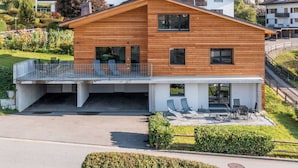 Una moderna casa a due piani in legno con un balcone, un portico coperto e vista sugli edifici circostanti.