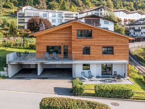 Ein modernes, zweistöckiges Holzhaus mit Balkon, überdachter Terrasse und Blick auf die umliegenden Gebäude.