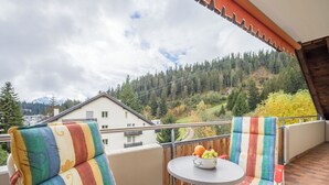Ein Balkon mit zwei gestreiften Stühlen, einem kleinen runden Tisch mit Früchten und Blick auf einen bewaldeten Hang.