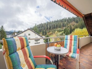 Un balcón con dos sillas a rayas, una mesa redonda pequeña con fruta y vistas a una ladera boscosa.