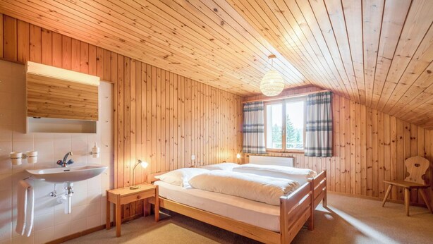 Ein Schlafzimmer mit Holzwänden, einem Bett, einem Nachttisch, einer Lampe, einem Fenster mit Vorhängen und einem Waschbecken.