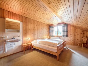 Un dormitorio con paneles de madera, una cama, una mesita de noche, una lámpara, una ventana con cortinas y un lavamanos.