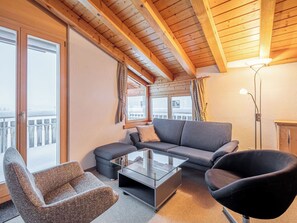 Een moderne woonkamer met een bank, fauteuil en salontafel, houten balken in het plafond en grote ramen met gordijnen.