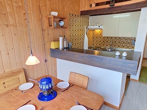 A wooden interior with a dining table, chairs, a blue electric kettle, a wall-mounted shelf with kitchen items, and a kitchen area with a stove and sink. - Beautiful apartment for 4 guests with WIFI, TV, terrace, pets allowed and panoramic view (Sagogn)