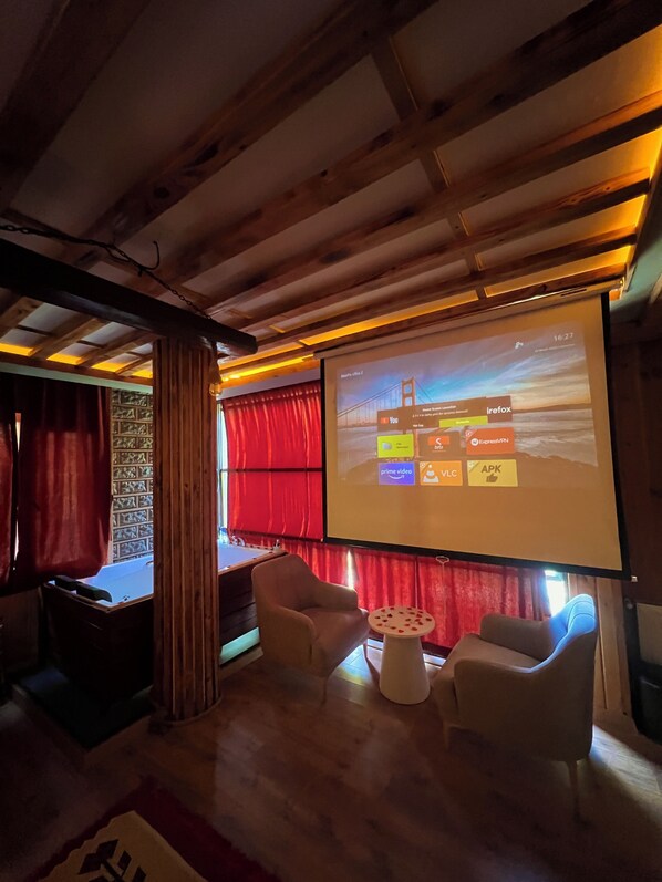Una habitación acogedora con techo de madera, una pantalla de proyector mostrando una presentación, dos sillones blancos y una pequeña mesa redonda con un centro de mesa de flores rojas.