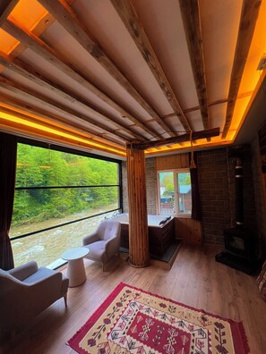 A cabin interior with a large window, wooden ceiling beams, a fireplace, and a cozy seating area. - VALLEY SUİT (Çamlıhemşin)