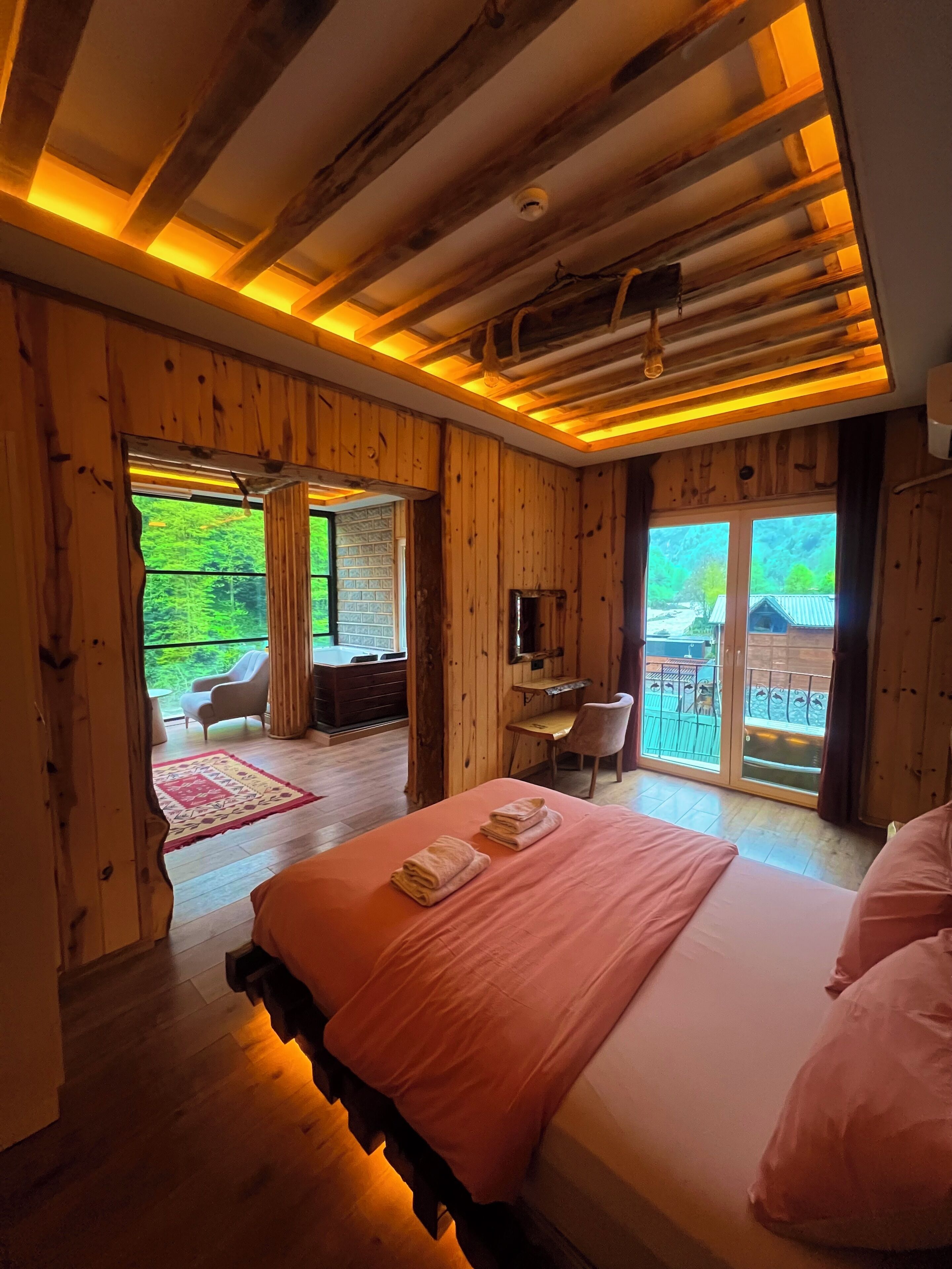 A wooden room with a bed, a desk, and a view of greenery outside.