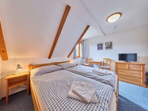 Ein Schlafzimmer mit schrägen Wänden, einem Bett mit karierter Bettwäsche, einem Schreibtisch mit Lampe, einem Sessel, einer Kommode und einem Fernseher.