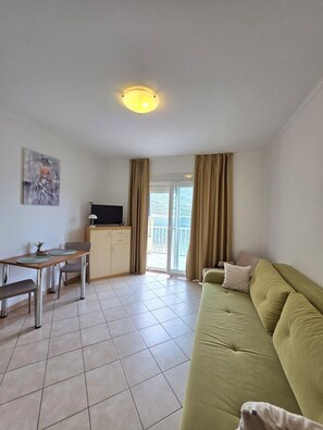 Uma sala de estar com um sofá verde, uma mesa de jantar com cadeiras, uma TV em um armário e uma varanda com cortinas.