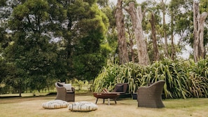 An outdoor seating area with wicker furniture and cushions, surrounded by tall trees and greenery. - Istana Lodge - 5 Bed house on acreage with pool (Musk Vale)
