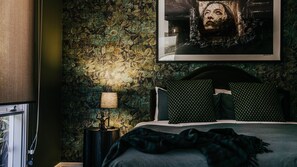 Ein Schlafzimmer mit strukturierter Wand, einem Bett mit dunkler Bettwäsche, zwei gemusterten Kissen, einem Nachttisch mit Lampe und einem gerahmten Kunstwerk über dem Bett.