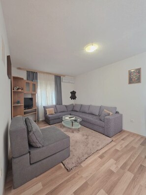 Uma sala de estar com um sofá modular cinzento, uma mesa de centro de vidro, um móvel para TV de madeira e um tapete em tons claros.