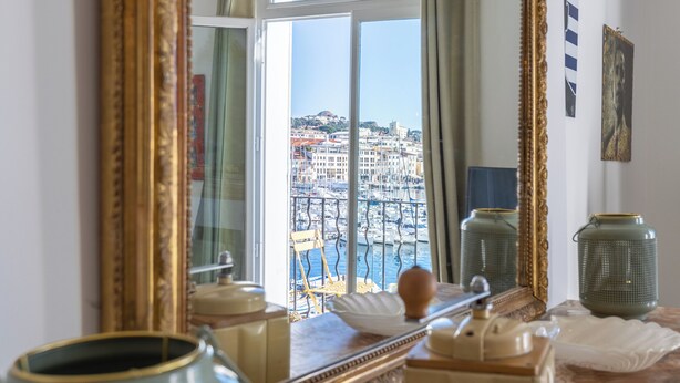 Ein Zimmer mit einem großen Spiegel, der einen Blick auf eine Stadtlandschaft reflektiert, einem Marmorboden und einem Fenster mit Vorhängen.