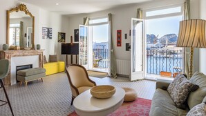 Ein großzügiger Wohnraum mit einer Couch, Sesseln, einem Hocker, einem Kamin, einem Fernseher und einem großen Fenster mit Blick auf eine Brücke und Wasser.