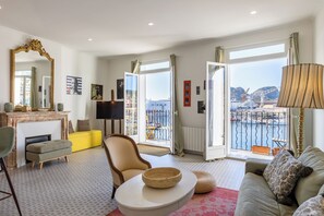 A spacious living room with a sofa, armchair, and ottoman, a fireplace, a television, and a large window with a view of a bridge and water. - The "Classé VIP", a balcony perched on the Old Port (La Ciotat)