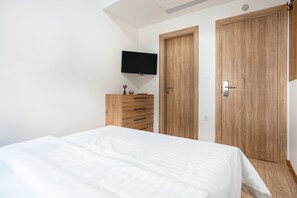 Quarto com cama, cómoda de madeira, televisão fixada na parede e duas portas de madeira.