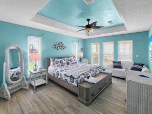 A bedroom with a bed, a chair, a nightstand, a ceiling fan, and a window with blinds. - Pelican and Palms 3 bed 2.5 baths Sleeps 14 Ocean View 3 minute walk to beach (Crystal Beach)