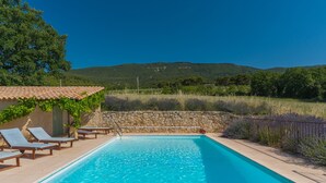 A swimming pool with lounge chairs, a stone wall, and a view of hills.