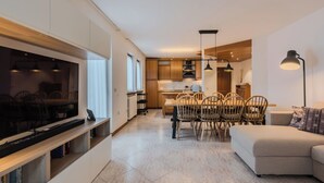 Un soggiorno moderno con una TV a schermo piatto, una zona pranzo con sedie di legno, una cucina con mobili in legno e un divano.